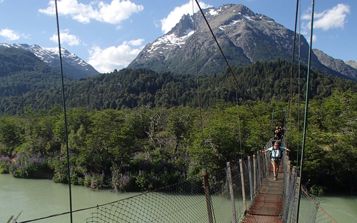 A group of people make their way over a swing bridge that hangs over a river. In the background, there are thick green trees in front of a mountain. 